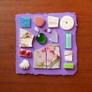 May include: A purple paper background with a variety of small, colorful, and patterned objects glued to it. The objects include a seashell, a green glass bead, a pink and white floral design, a blue and white floral design, a blue square, a red and white square, a white circle, a green square, a blue rectangle, a white rectangle, a green rectangle, and a pink and white floral design.