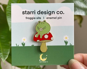 May include: Enamel pin featuring a green frog sitting on a red mushroom with white spots. The pin has a gold-colored stem and a small white flower. The pin is on a card with the text "starri design co." and "froggie sits | enamel pin."