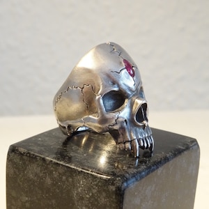 Könnte beinhalten: Ein silberner Totenkopfring mit einem roten Edelstein, der in die Stirn eingefasst ist. Der Ring ist mit Rissen und Spalten am Schädel verziert.