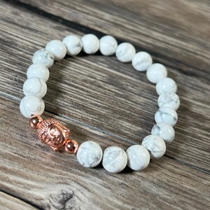 Könnte beinhalten: Weißes Howlith-Perlenarmband mit einem roségoldenen Buddha-Anhänger. Das Armband besteht aus natürlichen Steinen und ist eine großartige Möglichkeit, Ihrem Look einen Hauch von Stil und Spiritualität zu verleihen.