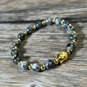 Könnte beinhalten: Ein graues und goldenes Perlenarmband mit einem goldenen Buddha-Kopf-Anhänger. Das Armband besteht aus runden Perlen und ist auf einer dehnbaren Schnur aufgefädelt.