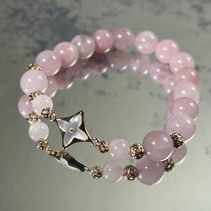 Könnte beinhalten: Ein Armband aus rosafarbenen Rosenquarzperlen mit einem goldfarbenen Sternanhänger. Der Anhänger hat eine weiße Perlmuttmitte.