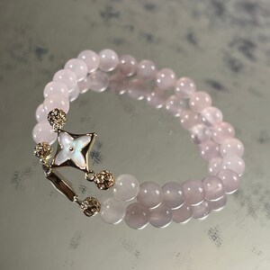 Könnte beinhalten: Ein Armband mit zwei Strängen aus hellrosa Perlen. Das Armband hat einen goldfarbenen Verschluss mit einem weißen, blütenförmigen Inlay und hängenden goldfarbenen Akzenten. Die Perlen sind glatt und rund, was einen zarten und eleganten Look erzeugt.