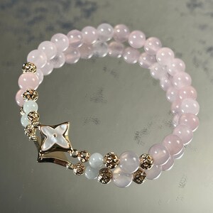 Könnte beinhalten: Ein rosa-weißes Perlenarmband mit einem goldenen Blumenanhänger. Das Armband besteht aus Rosenquarz- und weißen Jadeperlen.