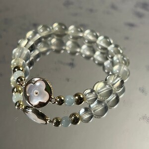 Könnte beinhalten: Ein Armband mit doppelter Schnur aus klaren und hellblauen Perlen, goldenen Akzenten und einem weißen Blumenanhänger.