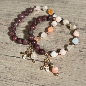 Könnte beinhalten: Zwei Armbänder mit Edelsteinperlen und goldenen Bienenanhängern. Die Armbänder sind aus rosa, weißen und orangefarbenen Perlen gefertigt, und die Anhänger sind mit kleinen Strasssteinen verziert.