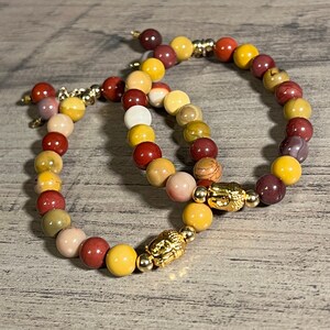 Könnte beinhalten: Zwei Armbänder aus bunten Steinperlen mit goldenen Akzenten. Jedes Armband hat einen goldenen Buddha-Anhänger.