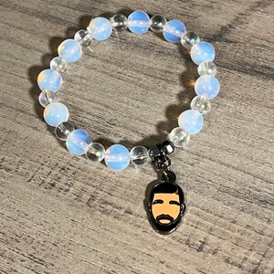 Könnte beinhalten: Ein Armband aus weißen Opalperlen mit einem silbernen Anhänger, der ein Cartoon-Bild eines Männergesichts zeigt.