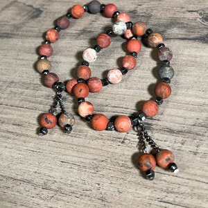 Könnte beinhalten: Zwei Armbänder aus roten, braunen und weißen Achatperlen mit schwarzen Hämatitperlen und silbernen Kettenquasten.
