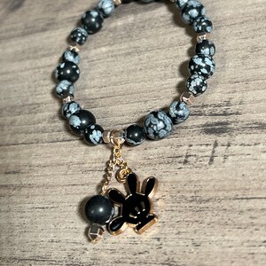 Könnte beinhalten: Ein Armband aus schwarzem und weißem Schneeflocken-Obsidian mit einem goldenen Anhänger in Form eines Comic-Kaninchens. Das Armband besteht aus runden Perlen und hat eine goldene Kette, die an dem Anhänger befestigt ist.