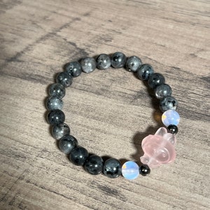 Könnte beinhalten: Ein schwarzes Perlenarmband mit einem rosa und weißen Kristall-Anhänger. Das Armband besteht aus runden Perlen und hat ein elastisches Band.