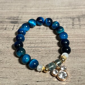 Könnte beinhalten: Ein blaues Tigerauge-Armband mit einem goldenen Anhänger mit drei klaren Strasssteinen. Der Anhänger ist in Form einer Mickey-Maus-Kopf gestaltet.