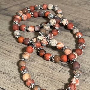 Könnte beinhalten: Drei rote, braune und weiße Stein-Armbänder mit silbernen Blumenakzenten. Die Armbänder sind übereinander gestapelt.