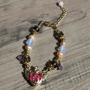 Könnte beinhalten: Ein goldenes Armband mit einem rosa Herz-Anhänger, klaren und undurchsichtigen Perlen und violetten Tropfen-Akzenten.