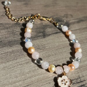 Könnte beinhalten: Ein zartes Armband aus rosa, weißen und goldenen Perlen. Das Armband hat einen goldenen Kettenverschluss und einen kleinen goldenen Blumenanhänger mit weißem Zentrum.