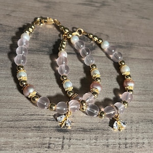 Könnte beinhalten: Zwei Armbänder mit Rosenquarz-, Perlen- und goldfarbenen Elementen. Jedes Armband hat einen kleinen, goldfarbenen Bienenanhänger. Die Armbänder liegen auf einer Holzoberfläche.