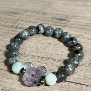 Könnte beinhalten: Ein Armband aus grauen und schwarzen Perlen mit einem lila Blumenanhänger und zwei weißen Perlen.