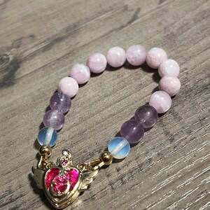 Könnte beinhalten: Ein Armband mit hellvioletten und Amethyst-Perlen, mit einem goldfarbenen Anhänger in Herzform mit Flügeln und rosa Akzenten. Das Armband liegt auf einer Holzoberfläche.