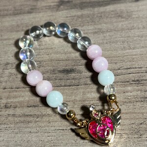 Könnte beinhalten: Ein rosa-weißes Perlenarmband mit einem goldenen Anhänger in Form eines rosa Herzens mit Flügeln. Der Anhänger hat einen kleinen rosa Stein in der Mitte.