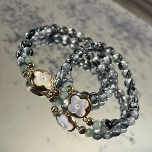 Könnte beinhalten: Zwei schwarze und silberne Perlenarmbänder mit goldenen Akzenten und weißen Blumenanhängern.