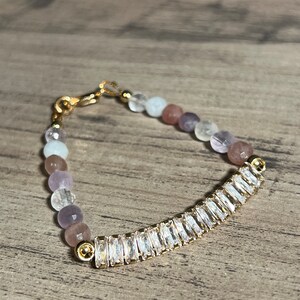 Könnte beinhalten: Ein zartes Armband mit einem goldfarbenen, gebogenen Stab mit klaren, rechteckigen Kristallen. Das Armband enthält auch eine Reihe kleiner, runder, mehrfarbiger Perlen in Rosa-, Lila- und Weißtönen. Das Armband wird mit einem goldenen Verschluss gesichert.