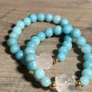 Könnte beinhalten: Zwei Armbänder aus hellblauen Perlen mit goldenen Akzenten und einem rosa geschnitzten Anhänger. Die Anhänger haben die Form einer Katze.