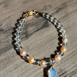 Könnte beinhalten: Ein Armband aus klaren Kristallperlen mit einem goldenen Verschluss und einem Anhänger aus hellblauem Edelstein. Das Armband hat abwechselnd klare und hellrosa Perlen.