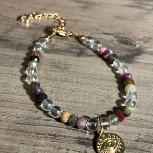 Könnte beinhalten: Ein goldenes Armband mit klaren Kristall- und mehrfarbigen Edelsteinperlen. Das Armband hat einen goldenen Anhänger mit einem Sonnen- und Augenmotiv.