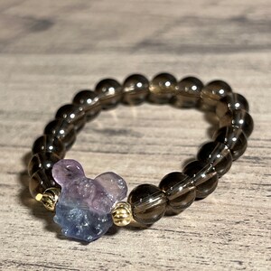 Könnte beinhalten: Ein dunkelbraunes Perlenarmband mit einem violetten und blauen Kristall-Anhänger. Der Anhänger ist in Form eines Mäusekopfes.