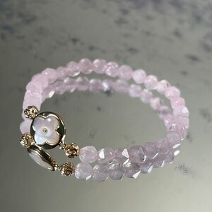 Könnte beinhalten: Ein doppelreihiges Armband aus hellrosa Perlen mit einem goldenen Blumenanhänger. Der Blumenanhänger hat eine weiße Mitte.