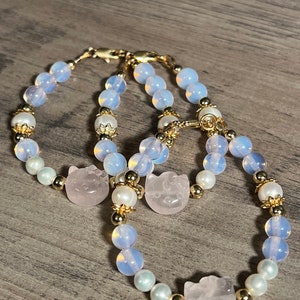Könnte beinhalten: Zwei Armbänder mit hellblauen und weißen Perlen, goldenen Akzenten und rosa Katzenanhängern.