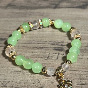 Könnte beinhalten: Ein grünes und rosafarbenes Perlenarmband mit einem goldenen Anhänger, der ein mehrfarbiges Auge zeigt. Das Armband hat goldene Abstandshalter zwischen den Perlen.