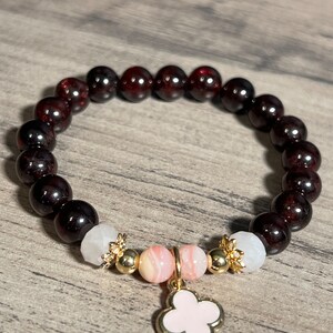 Könnte beinhalten: Ein Armband aus roten Edelsteinen mit einem rosa und weißen Anhänger. Der Anhänger ist in Form eines vierblättrigen Kleeblatts.