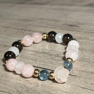 Könnte beinhalten: Ein Armband aus rosafarbenen, braunen und blauen Edelsteinperlen und goldenen Zwischenstücken. Das Armband hat einen rosenförmigen Anhänger.