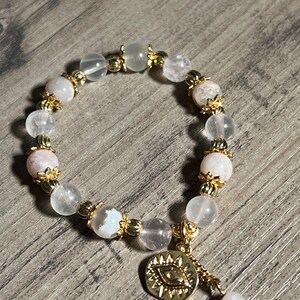 Könnte beinhalten: Ein weißes und rosafarbenes Kristall-Armband mit goldenen Akzenten und einem goldenen Anhänger mit einem bösen Auge.