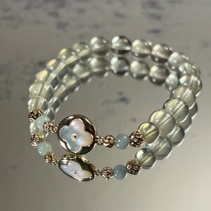 Könnte beinhalten: Doppelreihiges Armband mit transparenten, runden Perlen und goldfarbenen Metallakzenten. Das Armband hat ein zentrales Dekorationselement mit einem Kleeblattmuster aus Perlmutt und kleinen, hellblauen Perlen.