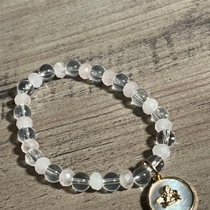 Könnte beinhalten: Ein Armband aus klaren und rosafarbenen Kristallperlen mit einem goldenen Anhänger in Form eines Engels. Der Anhänger ist an einem weißen Perlmutt-Hintergrund befestigt.