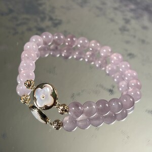 Könnte beinhalten: Ein Armband mit zwei Reihen aus helllila Perlen und einem goldenen Anhänger mit einem weißen Blumenmotiv.