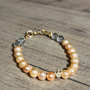 Könnte beinhalten: Ein zartes Armband mit einer Mischung aus klaren und pfirsichfarbenen Perlen und einem kleinen goldenen Kronenanhänger.