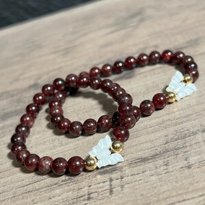 Könnte beinhalten: Zwei rote Edelstein-Armbänder mit goldenen Akzenten und weißen Schmetterlingsanhängern.