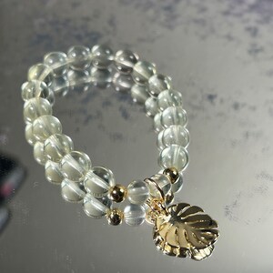 Könnte beinhalten: Ein Armband aus klarem Quarz mit goldenen Perlen und einem goldenen Blattanhänger. Das Armband besteht aus kleinen, runden Perlen und ist auf einer durchsichtigen Gummikordel aufgefädelt.