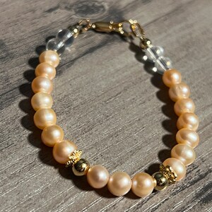 Könnte beinhalten: Ein Armband aus hellrosa Perlen mit goldenen Akzenten. Das Armband hat einen durchsichtigen Verschluss.