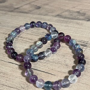 Könnte beinhalten: Zwei Armbänder aus runden Perlen in Lila, Blau und Klar. Die Perlen sind auf einem Gummiband aufgefädelt.