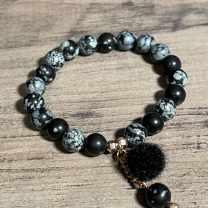 Könnte beinhalten: Schwarzes und weißes Schneeflocken-Obsidian-Armband mit einem schwarzen Bommel-Anhänger und einer goldenen Kette.