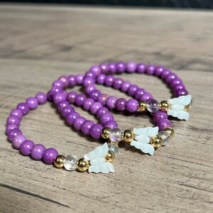 Könnte beinhalten: Drei violette Perlenarmbänder mit goldenen Akzenten und weißen Schmetterlingsanhängern.