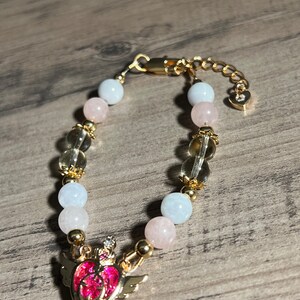 Könnte beinhalten: Ein goldenes Armband mit einem rosa Herz-Anhänger, verziert mit einer Krone und Flügeln. Das Armband besteht aus hellblauen, rosa und klaren Perlen.
