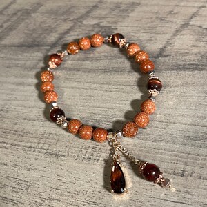 Könnte beinhalten: Ein braunes und goldenes Perlenarmband mit einem tropfenförmigen Anhänger. Das Armband besteht aus kleinen, runden Perlen und hat einen silbernen Verschluss.