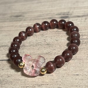 Könnte beinhalten: Ein braunes Perlenarmband mit einem rosa Blumenanhänger und goldenen Akzenten.