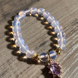 Könnte beinhalten: Ein Armband mit opalisierenden, runden Perlen. Es hat goldfarbene Akzente und einen Anhänger mit einem lila Edelstein, der von kleineren, klaren Steinen umgeben ist. Das Armband wird auf einer Holzoberfläche präsentiert.