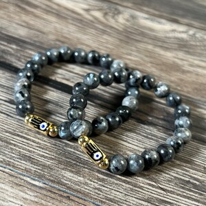 Könnte beinhalten: Zwei schwarze Perlenarmbänder mit goldenen Akzenten und blauen Glücksaugen-Anhängern. Die Armbänder liegen auf einer Holzoberfläche.